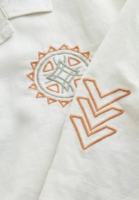 Valge kangas eristuva tikandiga: rohelise ja virsikuvärvi kolmnurkse aktsendiga ümmargune päikesemotiiv ning virsikuvärvi zigsagimustriga.