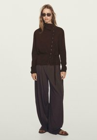 Pullover a maglia di colore marrone scuro con collo a imbuto, chiusura laterale con bottoni e vita legata. Abbinato a pantaloni larghi scuri e sandali flat.