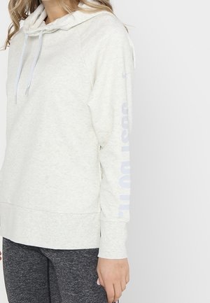 Frau trägt hellgrauen Hoodie mit Kordeln und dunkelgraue Leggings vor neutralem weißen Hintergrund.