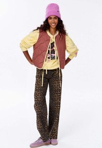 Hnědá prošívaná vesta přes žlutou mikinu s potiskem, doplněná leopardími kalhotami a fialovými teniskami, zakončená růžovou čepicí.