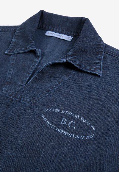 Chemise en denim bleu foncé avec col, avec un petit motif de texte ovale blanc brodé et une étiquette de la marque Bobo Choses à l'intérieur du col.