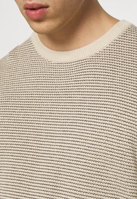 Detailní záběr na osobu s béžovým a černým pruhovaným pleteným svetrem s žebrovaným kulatým výstřihem.