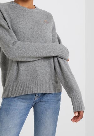 Personne portant un pull en tricot gris clair avec des poignets et un ourlet côtelés, associé à un jean bleu clair sur un fond blanc uni.