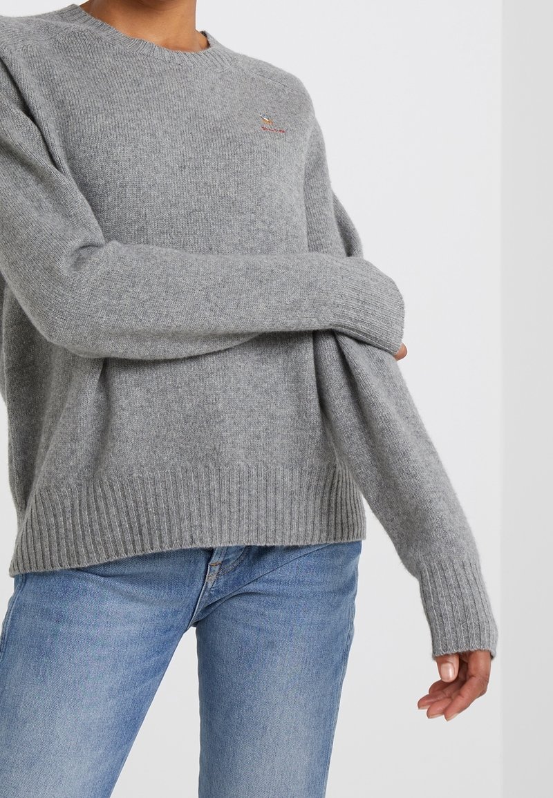 Personne portant un pull en tricot gris clair avec des poignets et un ourlet côtelés, associé à un jean bleu clair sur un fond blanc uni.