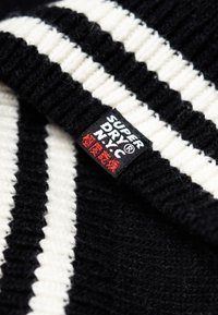 Tessuto a maglia bianco e nero con un ampio motivo a righe, caratterizzato da un'etichetta nera prominente con scritte rosse che recitano "Superdry N.Y.C.".