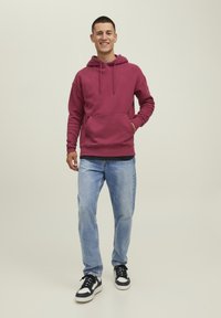 Felpa bordeaux con tasca anteriore e cordino regolabile, abbinata a jeans azzurri chiari e sneakers nere e bianche. Vestibilità e design casual.