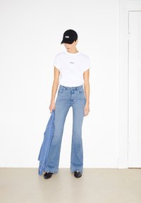 Vit kortärmad t-shirt, ljusblå utsvängda jeans, svart keps med "JA", håller en blå denimjacka. Slätt tyg och avslappnad passform.