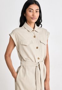 Μπεζ φόρεμα με κοντά μανίκια, δύο τσέπες στο στήθος και κουμπιά μπροστά. Διαθέτει ζώνη στη μέση και μαλακό, υφασμάτινο υλικό.