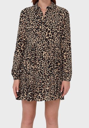 Vrouw die een luipaardprint jurk draagt met lange mouwen, knopen aan de voorkant, een laagjes rok en een kraag, staand tegen een effen achtergrond.