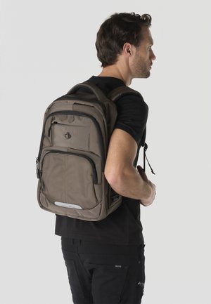 Hombre con camisa negra y mochila marrón con múltiples compartimentos con cremallera, de pie de perfil contra un fondo claro y liso.