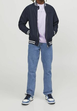 Person trägt eine marineblaue Collegejacke, ein helllila T-Shirt, blaue Jeans und weiße und blaue Turnschuhe und steht mit den Händen in den Taschen.