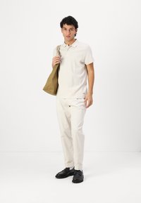 Polo shirt beige clair avec motif texturé, manches courtes, associé à un pantalon crème et des chaussures noires. Tenant un sac fourre-tout marron clair.