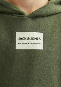 Sudadera verde con capucha y una textura suave que presenta una etiqueta rectangular blanca. La etiqueta tiene texto en negro: "JACK & JONES" y "Nuestro Legado, Tu Futuro".