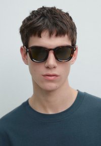 Occhiali da sole tartarugati con lenti scure, caratterizzati da una forma arrotondata e montature spesse. Indossati da una persona con una maglietta a girocollo scura.