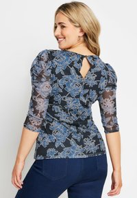 Blusa nera con maniche a sbuffo trasparenti, caratterizzata da motivi floreali blu. Dettaglio sulla schiena a chiave, orlo arrotondato e texture liscia.