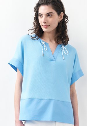 Frau mit lockigem braunem Haar, die ein locker sitzendes hellblaues Kurzarmshirt mit Kordelzug am Ausschnitt trägt und zur Seite schaut.