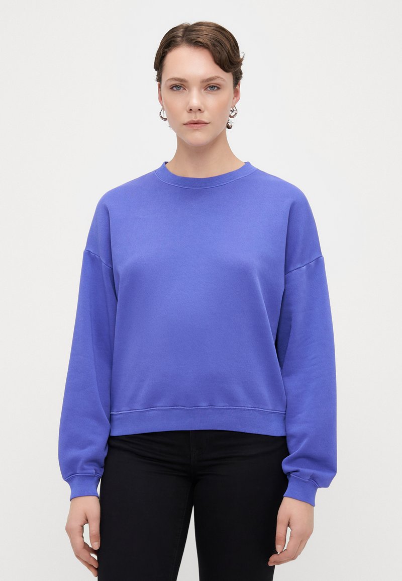 Sudadera de algodón morada con cuello redondo, hombros caídos y puños acanalados, combinada con pantalones negros, sobre un fondo liso.