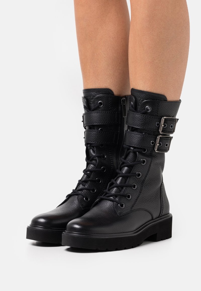 Bottes en cuir noires montant jusqu'au milieu du mollet, avec lacets, deux sangles à boucle ajustables, fermeture éclair latérale et semelle épaisse et robuste, portées sur des jambes nues.