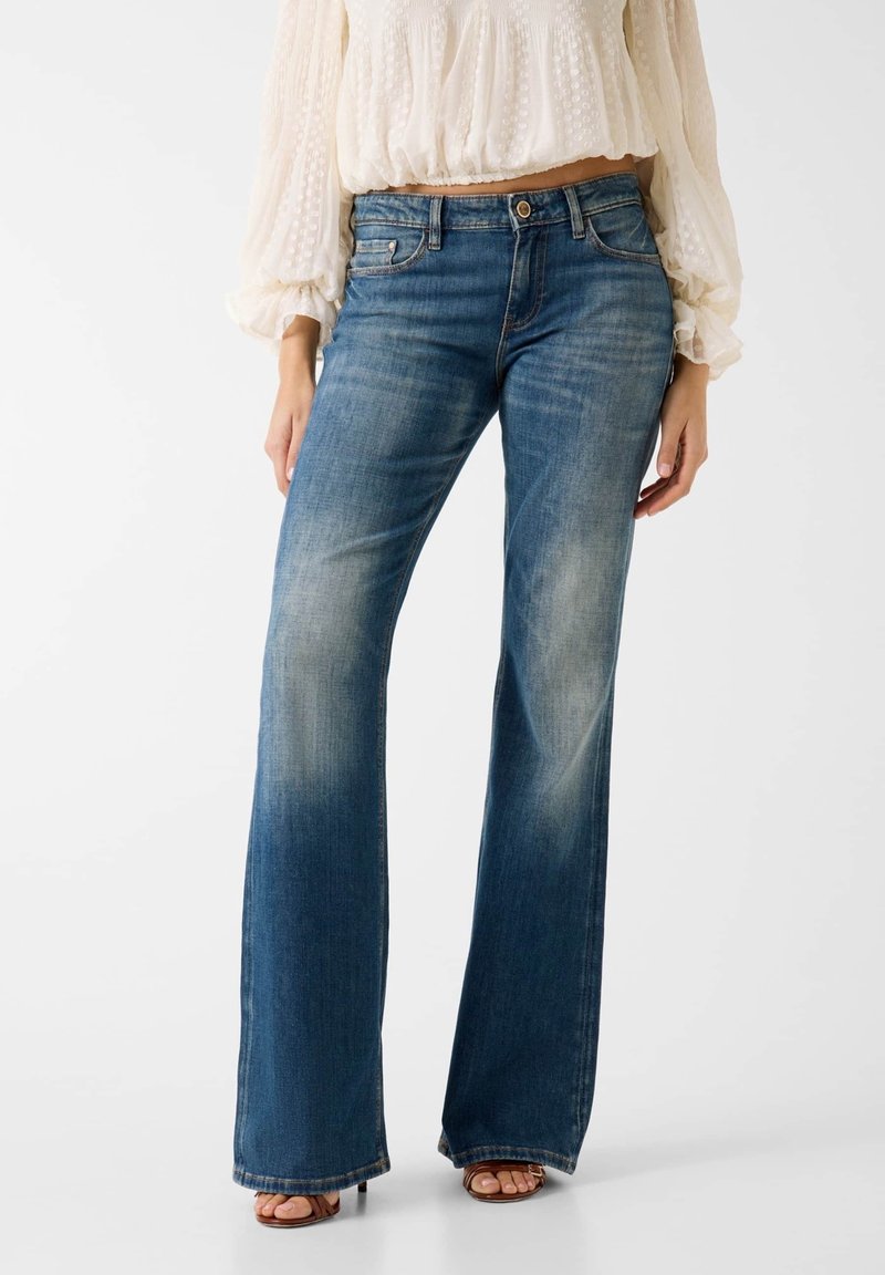 Femme portant un jean flare bleu délavé taille haute, associé à un chemisier crème texturé à manches longues et des sandales à bout ouvert.