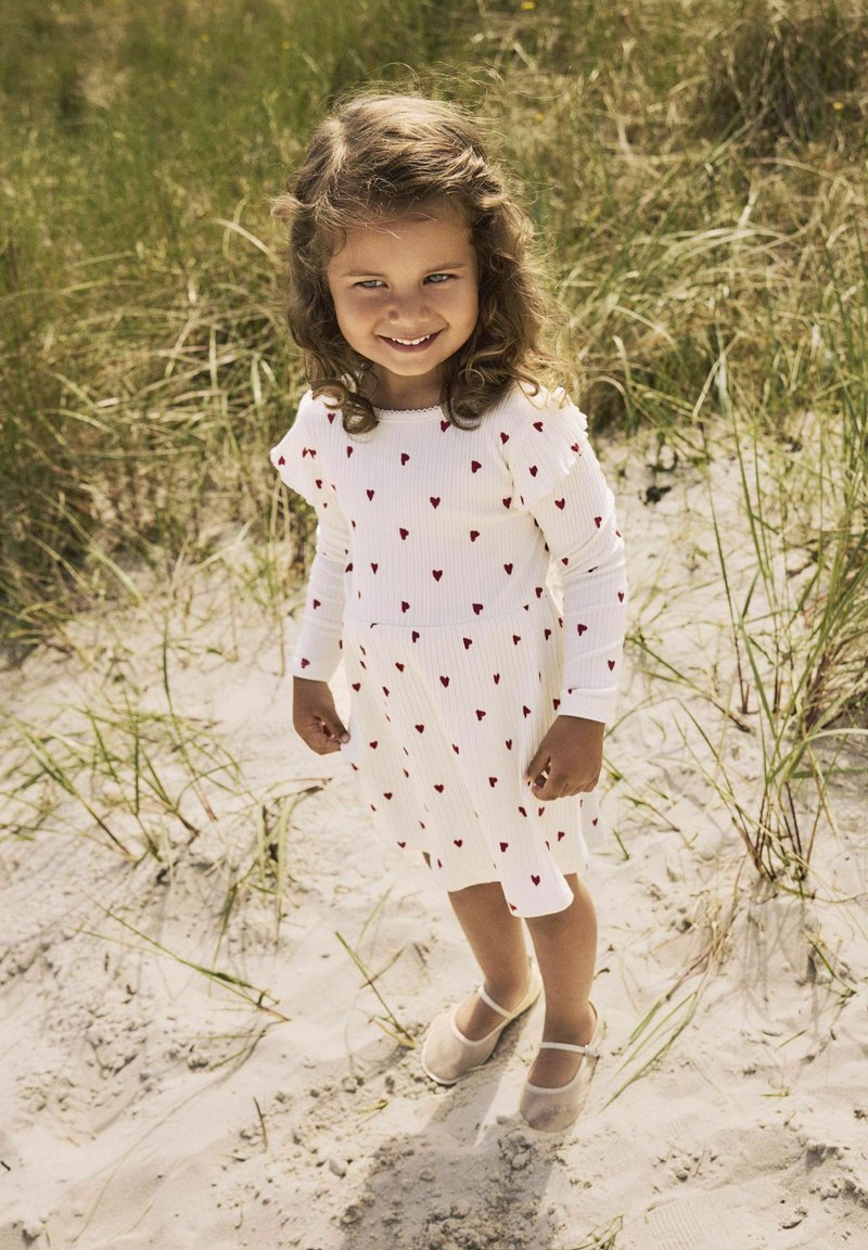 Robe blanche à rayures avec des motifs de cœurs rouges, manches longues et taille froncée. Chaussures claires avec des bandes. Fond sableux avec de l'herbe.