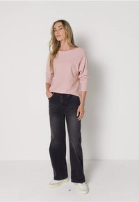 Frau trägt ein hellrosa langärmliges Oberteil, schwarze weite Jeans und weiße Sneakers und steht vor einem schlichten weißen Hintergrund.