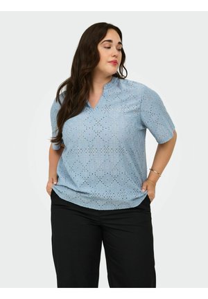 Kvinde med langt mørkt hår iført en lyseblå blonder kortærmet top og sorte bukser, hænderne i lommerne, kigger til venstre.