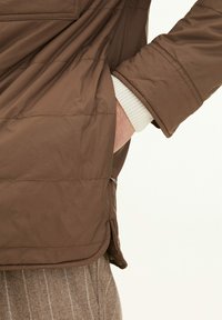 Giacca trapuntata marrone con la mano nella tasca laterale, polsini a maglia bianca e pantaloni in lana a righe marrone visibili dalla vista laterale.