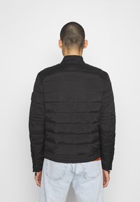 Black padded jacket with a stand collar, featuring horizontal stitch detailing and zippered cuffs, worn with light blue jeans.