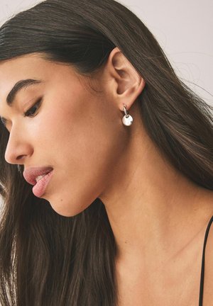 Woman with long dark hair wearing a silver hoop earring with a small round charm, facing left against a plain background.