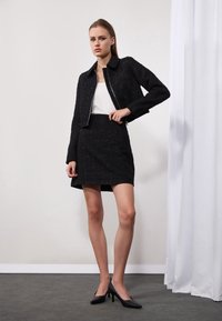 Svart tweed cropped jakke med glidelås og matchende skjørt, begge med et strukturert mønster. Modellen har på seg svarte høyhælte sko; hvit topp synlig under.
