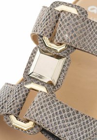 Beige sandalen met een gestructureerde band, metalen geometrische gesp en subtiele glans. Ontworpen voor comfort met een platte zool en open teen.