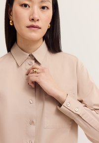 Beige Hemd mit Knopfverschluss, spitzem Kragen, einer einzelnen Brusttasche und glatter Textur. Verfügt über perlenartige Knöpfe vorne und an den Manschetten.