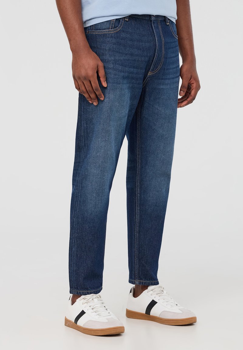 Jeans in denim blu scuro con taglio dritto, cinque tasche e dettagli di cucitura sottili. Indossati con sneakers bianche con accenti neri.