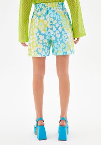 Kolorowe szorty z abstrakcyjnymi wzorami w odcieniach niebieskiego, zielonego i białego, z elastycznym pasem i luźnym krojem. Zestawione z niebieskimi sandałami na blokowym obcasie.