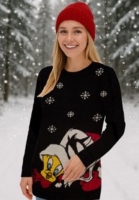 Maglione nero lavorato a maglia con fiocchi di neve e personaggi dei cartoni animati Tweety Bird e Sylvester. Una donna indossa un berretto di lana rosso e sorride.