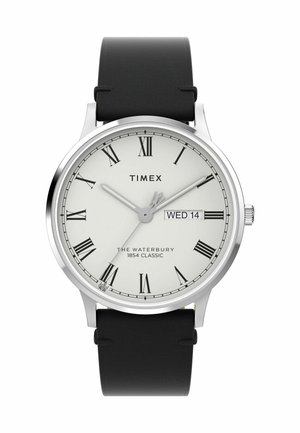La montre Timex Waterbury se caractérise par un boîtier rond en acier inoxydable argenté, un cadran blanc avec des chiffres romains, un bracelet en cuir noir et un affichage de la date.