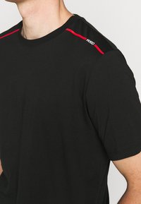 Sort bomulds T-shirt med korte ærmer, rund halsudskæring og røde accentstriber på skuldrene. Logo-detalje ved halsudskæringen.