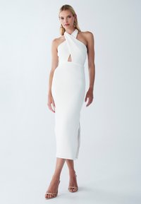 Weißes Halfterkleid mit einem Schlüsselloch-Ausschnitt, taillierter Silhouette, knielang, strukturierter Stoff und seitlichem Schlitz. Kombiniert mit nude-farbenen, schnürbaren High Heels.