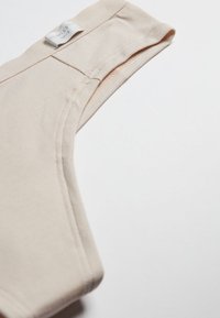 Béžové bezrukávové bavlněné tílko ukazující horní rameno a výstřih s viditelnou značkovou štítkem.