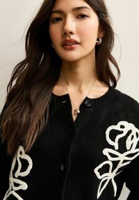 Femme aux longs cheveux foncés portant des boucles d'oreilles dorées en forme d'anneau, un collier avec pendentif, et un cardigan noir boutonné avec des motifs floraux blancs.