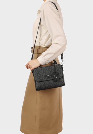 Donna che indossa una blusa e una gonna beige, che tiene una borsa a tracolla nera con motivo, dotata di tracolla regolabile e targhetta con logo.