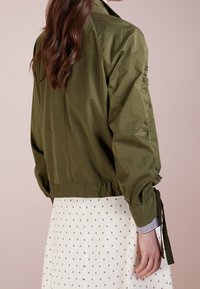 Olivgrön, lätt bomberjacka med samlade ärmar och justerbara snören. Matchad med en vit kjol med svarta stjärnmönster.