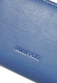 Portafoglio in pelle blu con superficie testurizzata. Presenta il logo "MAITRE" in rilievo, una cerniera liscia e un design rettangolare compatto.