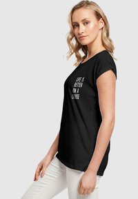 Sort bomulds t-shirt med korte rullede ærmer og hvid tekst, der står "LIVET ER BEDRE PÅ EN KRYDSTOGT." Model iført lyse bukser.