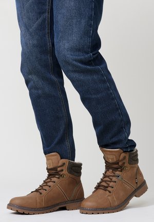 Botas de cuero marrón hasta el tobillo con un cuello texturizado, cordones y acentos metálicos. Combinadas con jeans de mezclilla oscura.