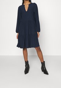 Robe bleu marine à manches longues, décolleté en V et jupe à volants, en tissu texturé avec des embellissements subtils. Associée à des bottines noires.