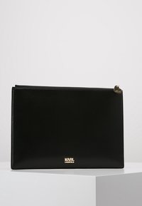 Musta nahkainen pussukka vetoketjulla, jossa kultainen logokuvio ja pieni kultainen koriste vetoketjussa. Sileä pinta ja suorakaiteen muoto.