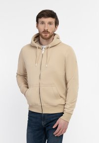 Beige zip-up hoodie med snoretræk, kengurulomme og ribbet mansjetter, båret over en hvid skjorte, parret med blå jeans.