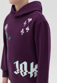 Sudadera morada de mezcla de algodón, con estampados gráficos en blanco y verde y capucha con cordones. El diseño incluye texto y patrones estilizados.