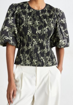 Kvinde iført en sort bluse med beige blomsterprint og puffede korte ærmer kombineret med højtaljede hvide bukser, hænder i lommerne.